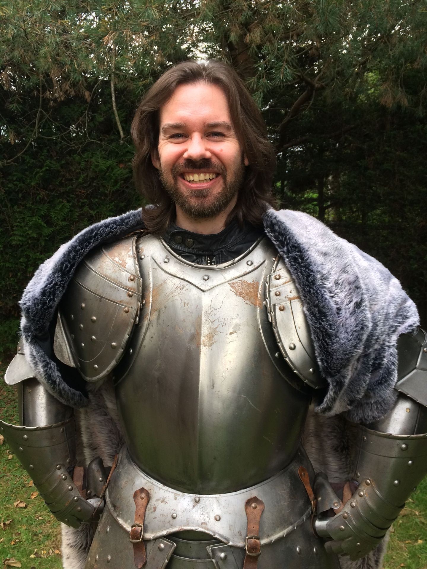 Person smiling in medieval armor with fur cloak, standing outdoors in front of trees.