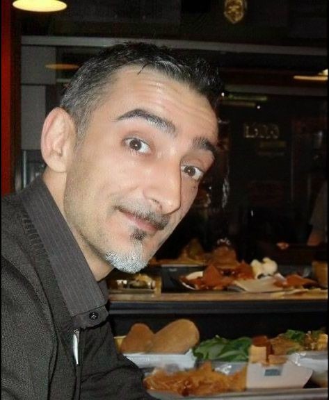 Man smiling with a background of various foods on a table in a restaurant setting.